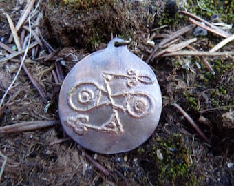 Puede incluir: Un colgante circular de metal con un acabado plateado deslustrado que presenta un diseño en relieve de dos círculos interconectados y símbolos estilizados en forma de flecha. El colgante tiene una pequeña anilla en la parte superior.
