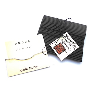 Op de afbeelding: Een witte kaart met het woord "AMOUR" in zwarte tekst en het woord "Code Morse" in zwarte tekst. De kaart heeft ook een zilveren armband met een staafbedel. De armband ligt op de kaart. De kaart ligt op een zwart-wit gestreept oppervlak. Een klein wit label met de tekst "Handmade with ♥ Caromb" is aan het oppervlak bevestigd.
