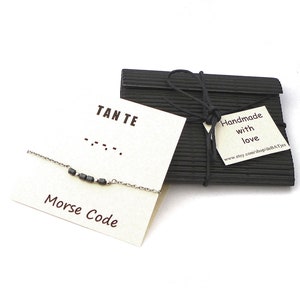 Puede incluir: Un brazalete de cadena plateada con cuentas negras que deletrean "TANTE" en código Morse. El brazalete se muestra en una tarjeta blanca con el texto "Código Morse" y "TANTE" en letras negras. La tarjeta está junto a una caja de regalo negra con una etiqueta que dice "Hecho a mano con amor" y una dirección de sitio web.