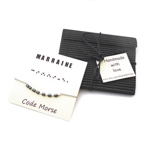 Peut inclure: Une carte blanche avec le mot "MARRAINE" écrit en lettres noires et une ligne de code Morse en dessous. La carte dit également "Code Morse". Une chaîne argentée avec des perles noires est sur la carte. Une boîte noire avec une étiquette blanche qui dit "Fait main avec amour" et une adresse de site Web est en arrière-plan.