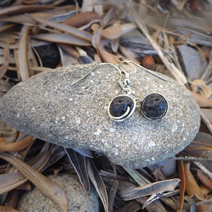 Op de afbeelding: Een paar zilveren oorbellen met zwarte lavastenen. De oorbellen hangen aan een steen.