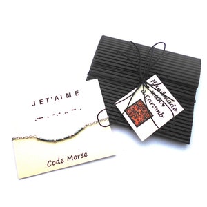 Op de afbeelding: Een zilveren kettingarmband met grijze kralen die "J'ET'AIME" in morsecode spellen. De armband is verpakt in een zwarte doos met een label waarop staat "Handmade with ♥ by Caromb".