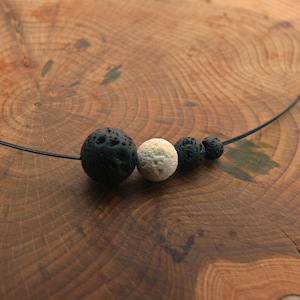 Op de afbeelding: Een ketting met zwarte lavasteen kralen en één crème-kleurige kraal. De kralen zijn geregen op een dun, zwart koord. De ketting wordt gepresenteerd op een houten oppervlak.