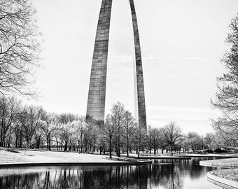 May include: A black and white photograph of the Gateway Arch in St. Louis, Missouri. The arch is reflected in a still body of water in front of it. Trees line the edge of the water.