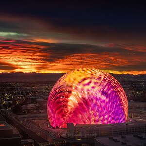 May include: A large, illuminated sphere with a pink and orange color scheme sits atop a building against a backdrop of a city skyline and a fiery sunset.