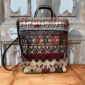 Op de afbeelding: Een veelkleurige, patroon tote bag met een zwart leren handvat en riem. De tas heeft verschillende geweven patronen in tinten rood, bruin en beige, geaccentueerd met zilverkleurige versieringen en blauwe kralen. De tas staat op een houten oppervlak.