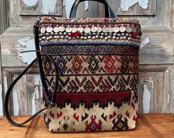 Puede incluir: Bolso tote estampado multicolor con asa y correa de cuero negro. El bolso presenta una variedad de patrones tejidos en tonos rojos, marrones y beige, acentuados con adornos plateados y cuentas azules. El bolso está sobre una superficie de madera.