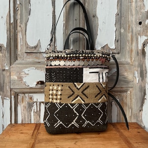 May include: A patchwork tote bag with a black leather handle and strap. The bag features a variety of patterned fabrics in black, white, and brown, with decorative metal accents along the top. The bag is sitting on a wooden surface.