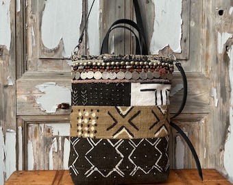Puede incluir: Un bolso tote patchwork con asa y correa de cuero negro. El bolso presenta una variedad de telas estampadas en negro, blanco y marrón, con detalles metálicos decorativos en la parte superior. El bolso está sobre una superficie de madera.