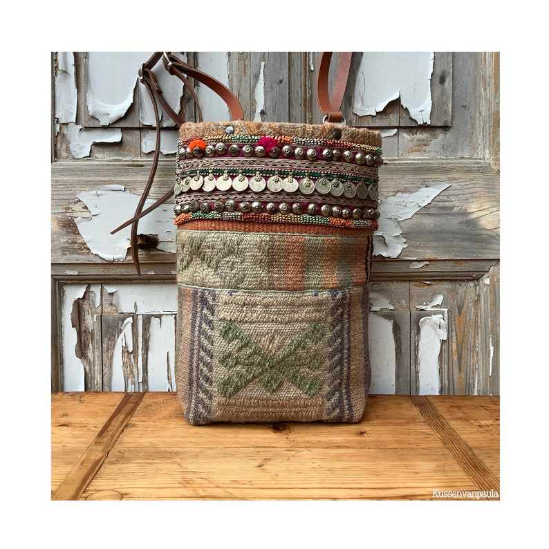 Puede incluir: Un bolso tejido marr&oacute;n con una correa larga. El bolso est&aacute; decorado con cuentas de colores, monedas de metal y borlas. El bolso est&aacute; sobre una superficie de madera.