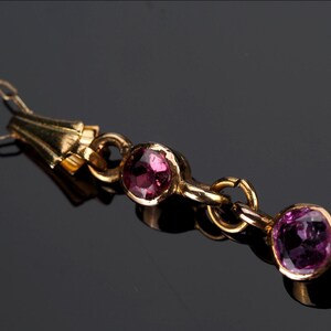 May include: A gold-toned necklace featuring a delicate chain and a series of circular settings, each containing a faceted, round, deep pink gemstone. The metal links connect the stones in a linear arrangement, creating a refined jewelry piece.
