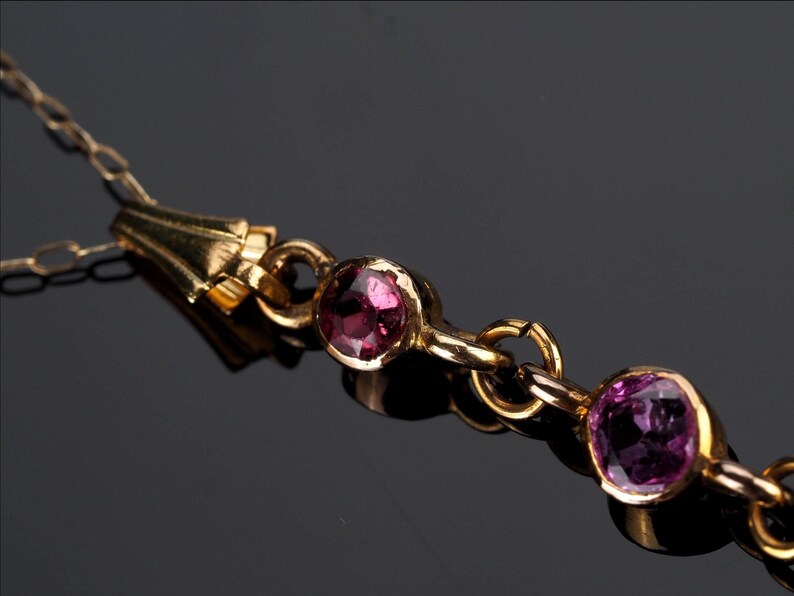 May include: A gold-toned necklace featuring a delicate chain and a series of circular settings, each containing a faceted, round, deep pink gemstone. The metal links connect the stones in a linear arrangement, creating a refined jewelry piece.