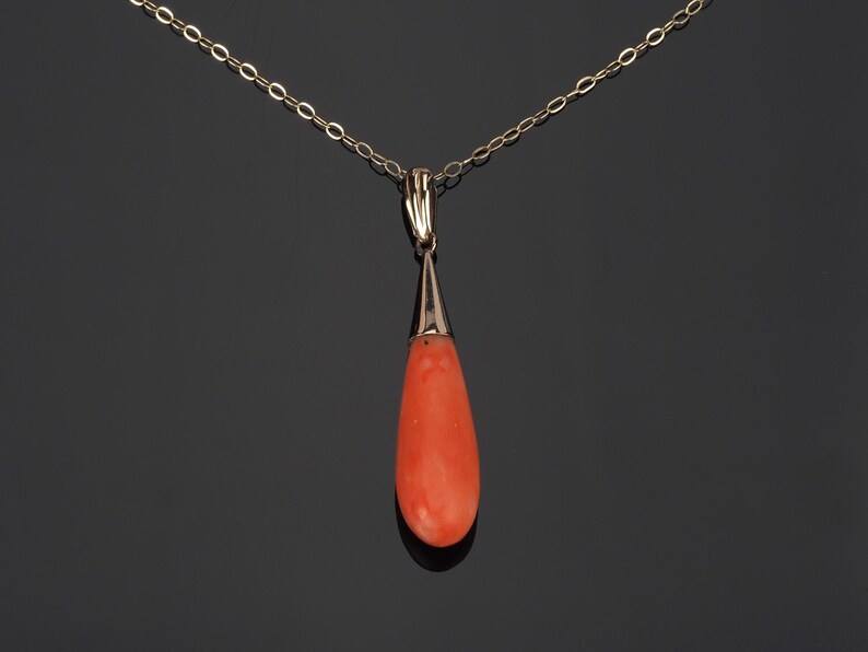 Pu&ograve; includere: Un ciondolo in corallo arancione a forma di goccia sospeso a una delicata catenina d'oro. La pietra di corallo liscia e lucida &egrave; incastonata in una semplice capsula d'oro che si collega alla catena a maglie sottili, creando un gioiello elegante.