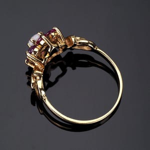 Puede incluir: Un anillo de oro con un grupo de piedras preciosas. El anillo presenta una piedra blanca central, flanqueada por piedras rojas oscuras m&aacute;s peque&ntilde;as. La banda y el engaste son de color oro pulido.
