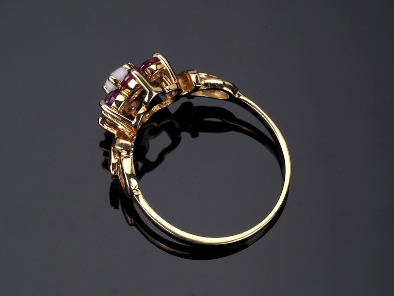 Puede incluir: Un anillo de oro con un grupo de piedras preciosas. El anillo presenta una piedra blanca central, flanqueada por piedras rojas oscuras m&aacute;s peque&ntilde;as. La banda y el engaste son de color oro pulido.