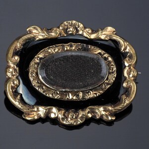 May include: An antique gold-toned brooch with an oval black center and a smaller oval inset. The brooch has an ornate, filigree-style frame with decorative flourishes. The background is dark, highlighting the brooch's details.