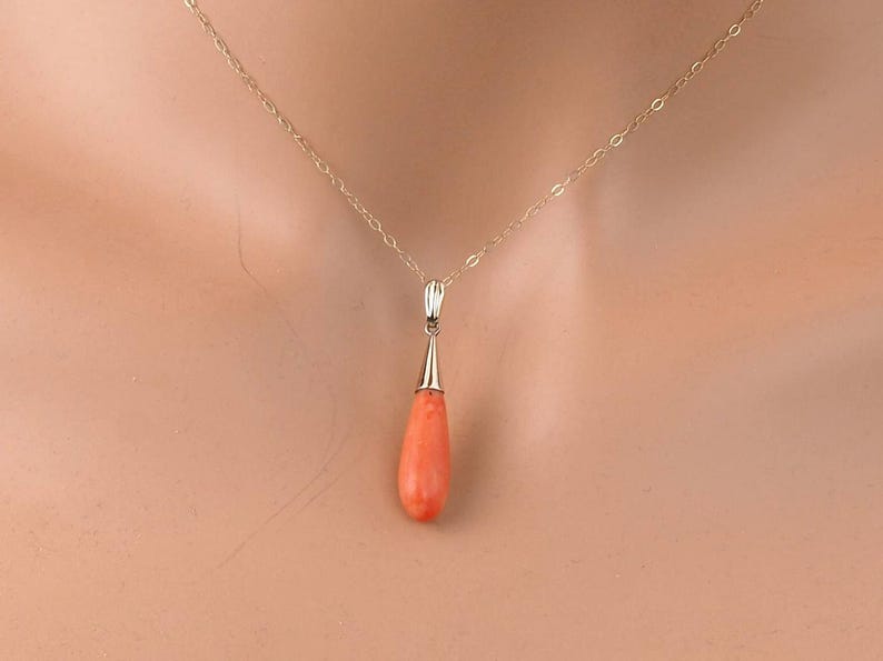 Pu&ograve; includere: Una collana a catena in oro con un ciondolo in corallo liscio a forma di goccia. Il ciondolo &egrave; fissato da un cappuccio in oro lucido sulla parte superiore, che si collega a una delicata catena a maglie d'oro attorno al collo.