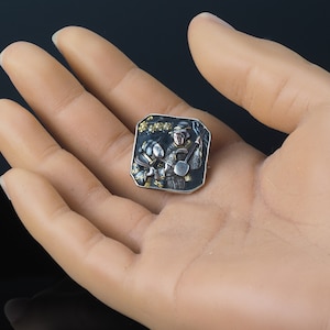 Pu&ograve; includere: Un fermaglio quadrato in argento con sfondo nero e dettagli dorati. Il fermaglio raffigura una scena di due figure che suonano strumenti musicali.