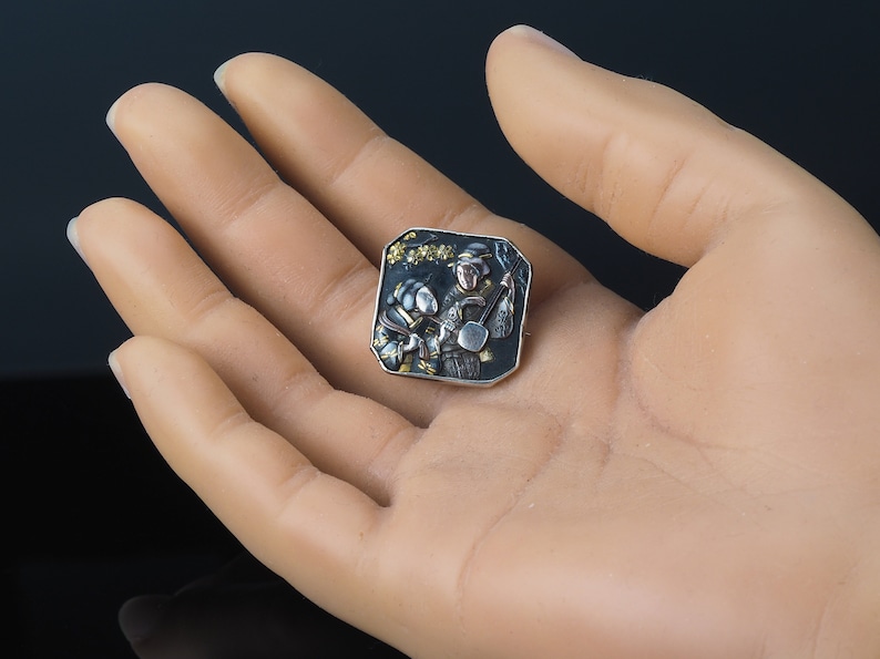 Pu&ograve; includere: Un fermaglio quadrato in argento con sfondo nero e dettagli dorati. Il fermaglio raffigura una scena di due figure che suonano strumenti musicali.