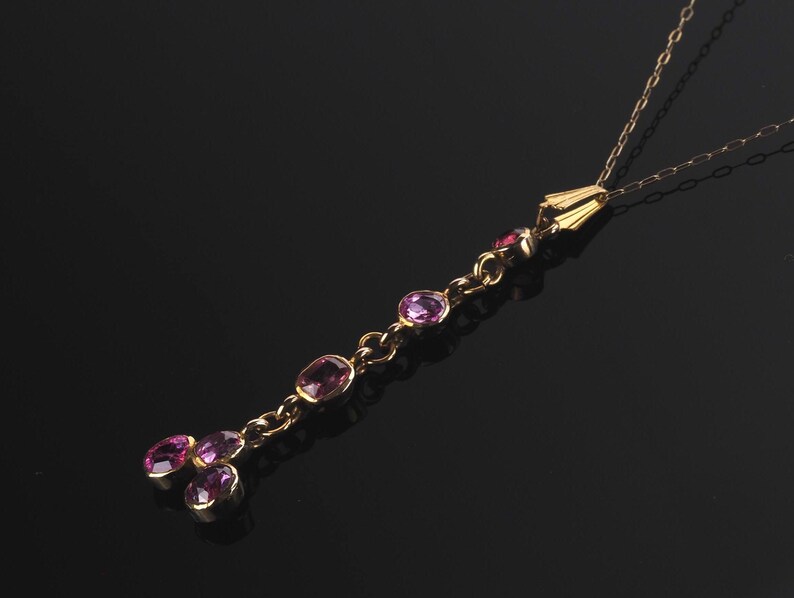 May include: A gold chain necklace featuring a vertical drop pendant set with five oval-cut purple gemstones. The pendant consists of a single line of four stones leading to a cluster of three stones at the base, all held in gold settings.