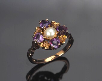 May include: A gold ring featuring a central white pearl surrounded by a cluster of four heart-shaped purple amethyst gemstones and four smaller heart-shaped yellow citrine gemstones arranged in a floral pattern on a polished gold band.