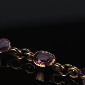 May include: A gold chain necklace featuring multiple bezel-set, cushion-cut purple gemstones. The links are rounded and connected to each stone setting, creating a continuous strand of metal and faceted violet-toned jewels.