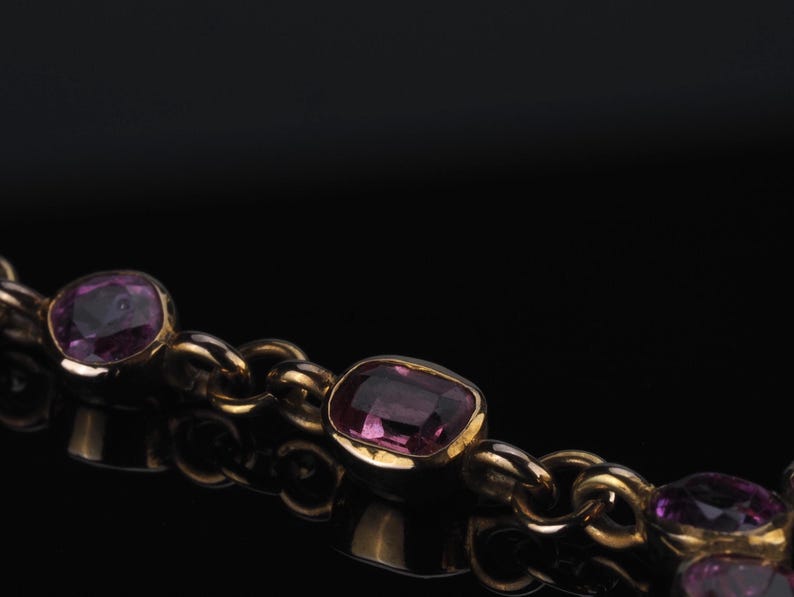 May include: A gold chain necklace featuring multiple bezel-set, cushion-cut purple gemstones. The links are rounded and connected to each stone setting, creating a continuous strand of metal and faceted violet-toned jewels.