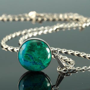 May include: A silver necklace with a round pendant featuring a vibrant teal and blue stone. The pendant is set in a silver bezel and hangs from a delicate chain. The necklace is displayed on a reflective surface.