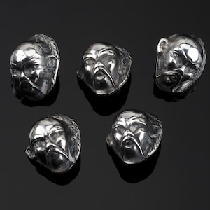 May include: Five silver-toned metal sculptures of stylized human faces with detailed features, including beards and facial expressions. The sculptures are arranged on a black surface, with each face appearing unique.