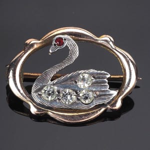 Puede incluir: Broche vintage de cisne con marco dorado. El cisne es plateado con tres gemas redondas y transparentes en su ala y un ojo de gema roja. El broche tiene forma ovalada.