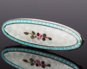 May include: An oval-shaped silver brooch with a white enamel background and a delicate floral design in pink, green, and brown. The brooch is outlined with a turquoise enamel border.