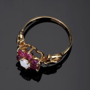 Puede incluir: Anillo de oro con dise&ntilde;o floral. Presenta una &oacute;palo blanco central rodeado de piedras rosas. La banda del anillo es dorada y la montura es detallada. Joyer&iacute;a.