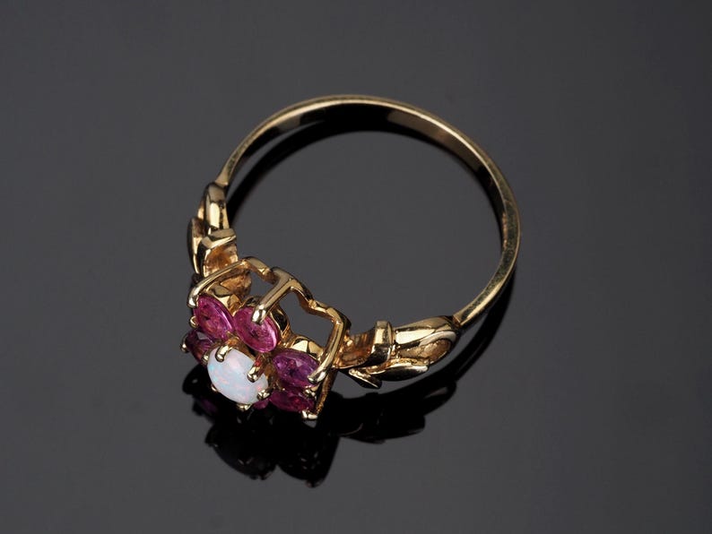 Puede incluir: Anillo de oro con dise&ntilde;o floral. Presenta una &oacute;palo blanco central rodeado de piedras rosas. La banda del anillo es dorada y la montura es detallada. Joyer&iacute;a.