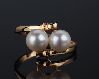 May include: A bypass style ring crafted from polished yellow gold, featuring two lustrous white pearls set at the open ends. The band has a textured, bamboo-like design that wraps around the finger, showcasing a reflective metallic finish.