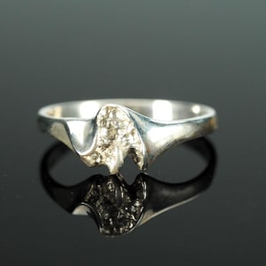 Puede incluir: Un anillo de plata con un diseño texturizado y abstracto en el centro. La banda del anillo se curva suavemente, y el diseño tiene una apariencia rugosa y orgánica. El anillo se muestra sobre una superficie negra reflectante.