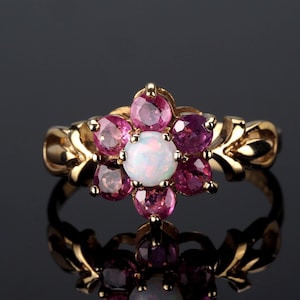 Puede incluir: Un anillo de oro con diseño floral, con una ópalo blanco central y gemas rosas y moradas circundantes. La banda del anillo tiene un patrón decorativo en forma de hoja.