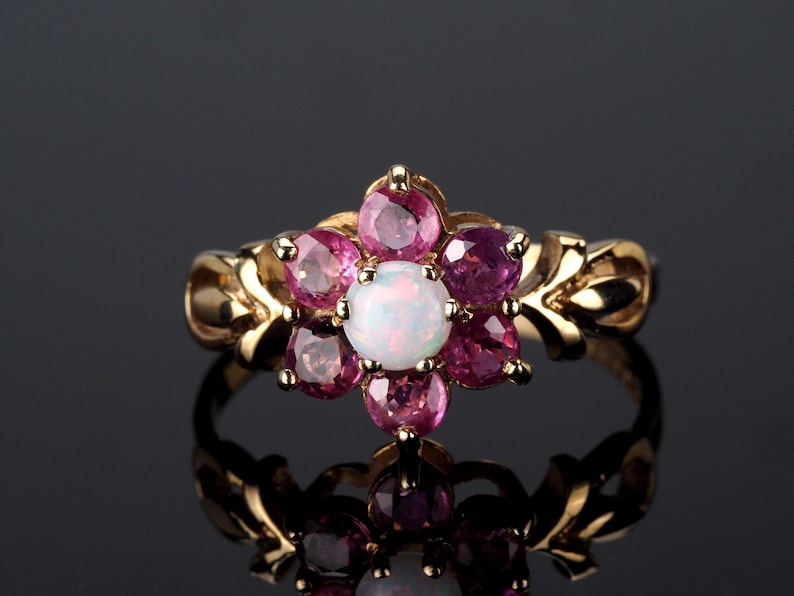 Puede incluir: Un anillo de oro con dise&ntilde;o floral, con una &oacute;palo blanco central y gemas rosas y moradas circundantes. La banda del anillo tiene un patr&oacute;n decorativo en forma de hoja.