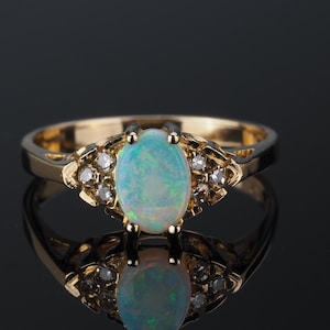 May include: A gold ring with an oval opal gemstone, displaying light blue and green hues. The ring is embellished with small diamonds. The ring is set against a black background.