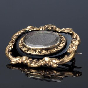 May include: An antique brooch with a gold-toned, ornate frame. The brooch features a black oval base with a clear, oval-shaped stone in the center. The gold frame has a detailed, swirling design.