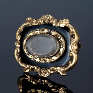 May include: Ornate gold-toned brooch with a dark blue enamel background. The brooch features an oval, possibly glass, center and intricate, scrolling details. The design is framed by a decorative gold border, creating a vintage aesthetic.