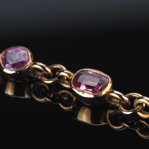 May include: A gold chain bracelet featuring multiple bezel-set, faceted pink gemstones. The links are circular and polished, connecting each individual stone setting in a continuous row. The metal has a warm, reflective yellow gold tone.