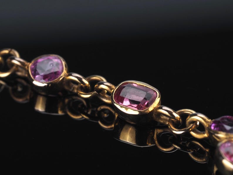 May include: A gold chain bracelet featuring multiple bezel-set, faceted pink gemstones. The links are circular and polished, connecting each individual stone setting in a continuous row. The metal has a warm, reflective yellow gold tone.