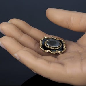 May include: An antique gold-toned brooch with a dark blue enamel background and a clear, oval-shaped stone. The brooch has an ornate, filigree-style frame. The brooch is held in an open hand.