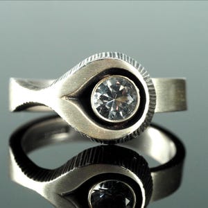 Puede incluir: Un anillo plateado con un engaste en forma de lágrima. El engaste tiene un diseño texturizado alrededor de una gema redonda y transparente. La banda es de diseño plano y sencillo. El anillo se muestra sobre una superficie reflectante.