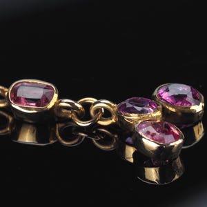 May include: A gold chain necklace featuring multiple bezel-set, faceted, deep pink oval gemstones. The stones are arranged in a cluster at the end of the chain, with one singular stone set further along the link, all set in polished yellow gold.
