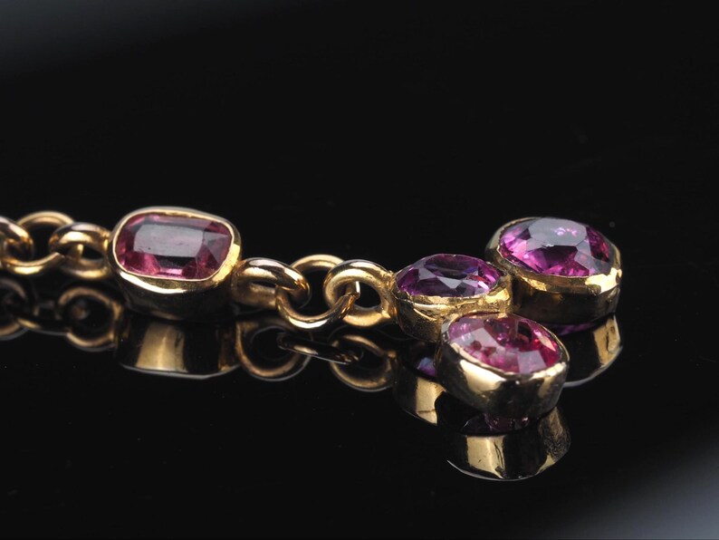 May include: A gold chain necklace featuring multiple bezel-set, faceted, deep pink oval gemstones. The stones are arranged in a cluster at the end of the chain, with one singular stone set further along the link, all set in polished yellow gold.