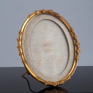 May include: An oval, gold-colored picture frame with a decorative floral design around the edge. The frame has a light-colored backing and a stand on the back to allow it to stand upright. The frame is empty.