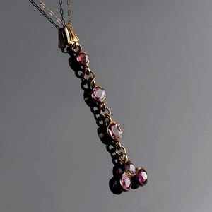 May include: A gold pendant necklace featuring a vertical chain of bezel-set pink gemstones. The design includes a series of individual stones connected by gold links, terminating in a cluster of three round pink gemstones at the bottom.