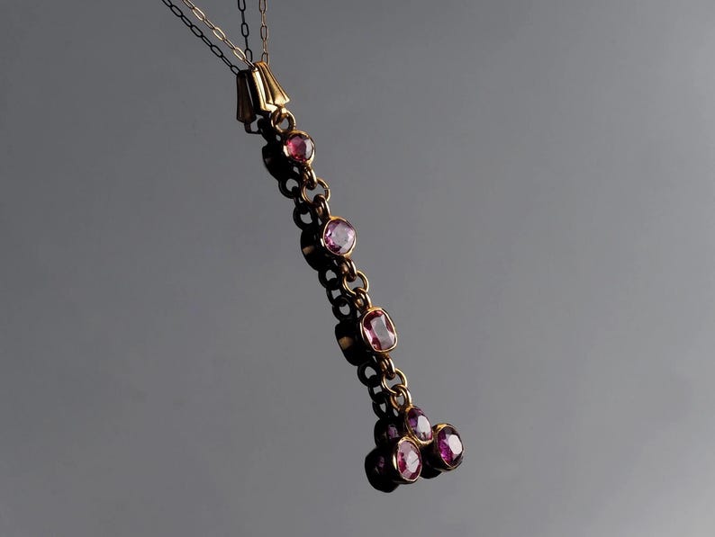 May include: A gold pendant necklace featuring a vertical chain of bezel-set pink gemstones. The design includes a series of individual stones connected by gold links, terminating in a cluster of three round pink gemstones at the bottom.
