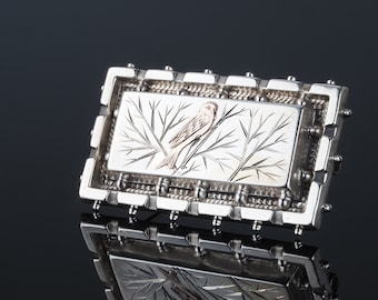 May include: A silver brooch with a detailed design of a bird perched on a branch. The bird is highlighted in gold.
